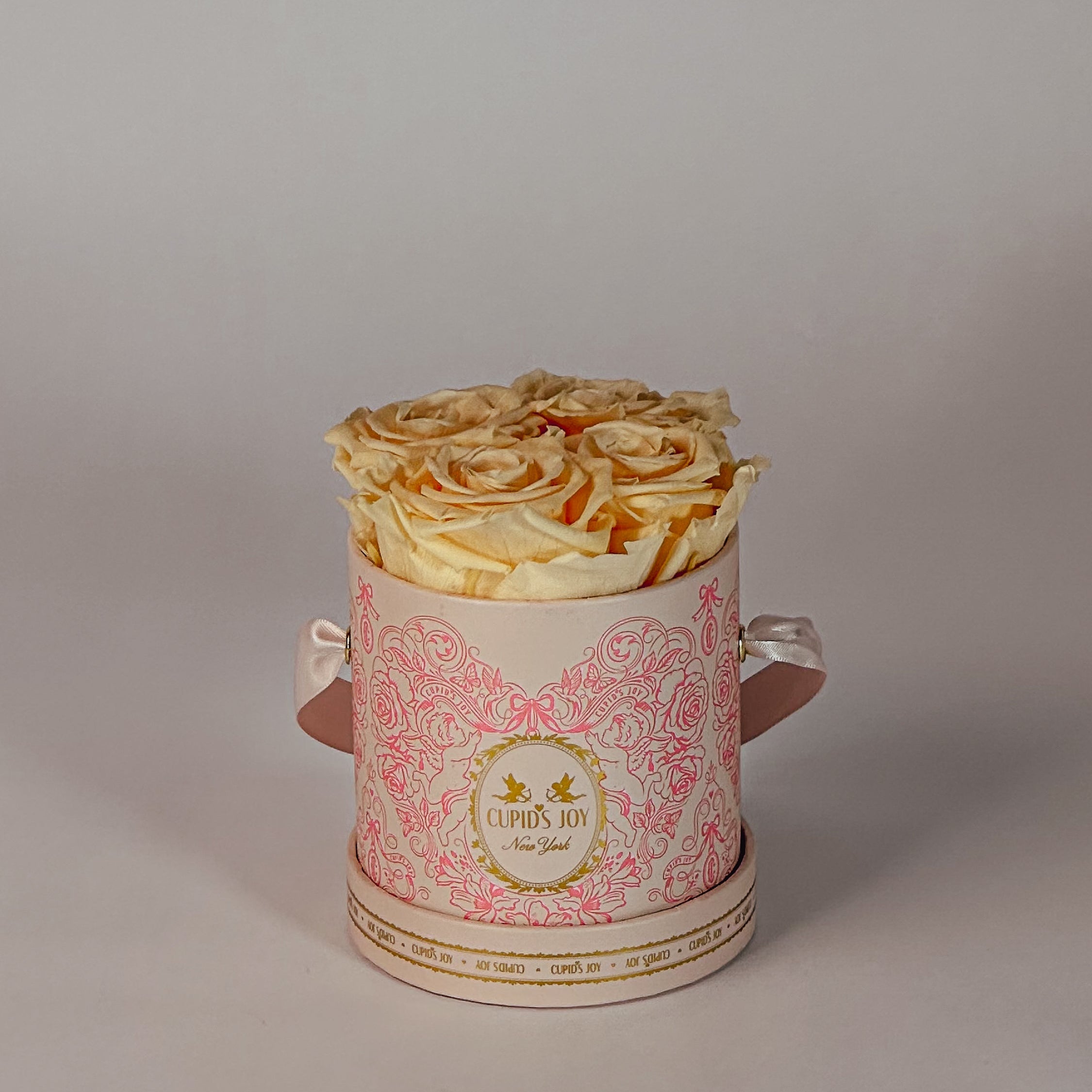 Decorative cupid-motif round box with 4 peach forever roses on a plain background