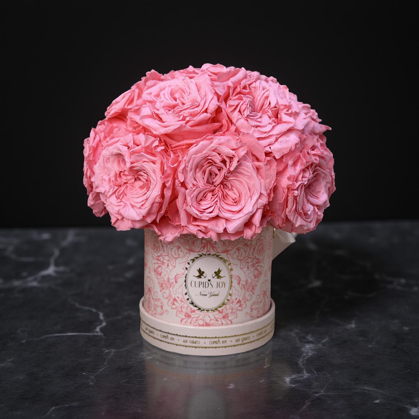 Pink eternal garden roses in a decorative round rose box on a dark marble surface with a black background
