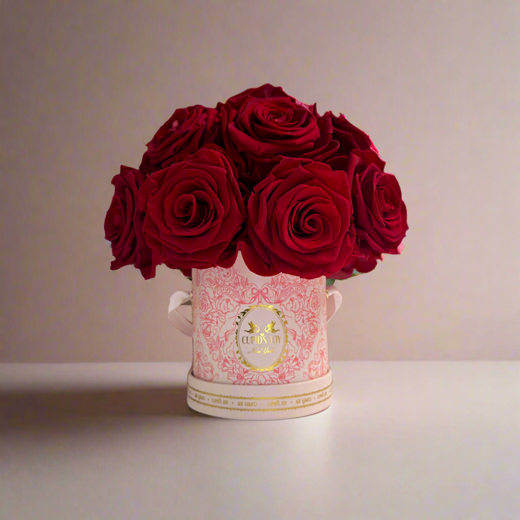 Arrangement of red eternal roses in a decorative pink and gold cupid-motif vase on a neutral background